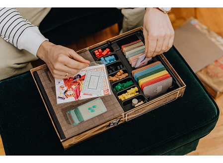 A person organises colourful cards, tokens, and game pieces inside a wooden board game box.